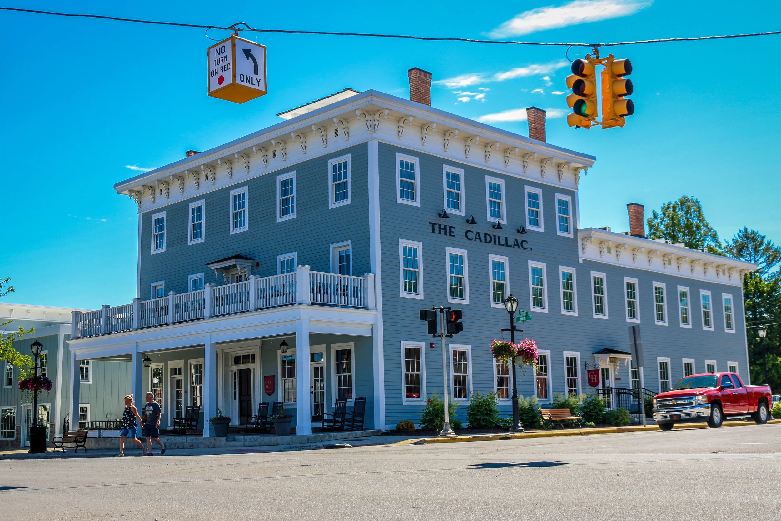 A large blue building called The Cadillac with multiple windows and white trim, situated on a street corner with a porch, potted plants, and hanging flower baskets. There are a few people walking, a red truck parked nearby, a traffic light, and street signs including one for a left turn only and a no turn on red.