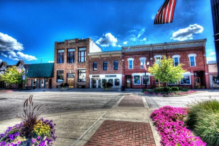 A small town main street with historic brick buildings, colorful flowerbeds, and an American flag, under a bright blue sky with scattered clouds.