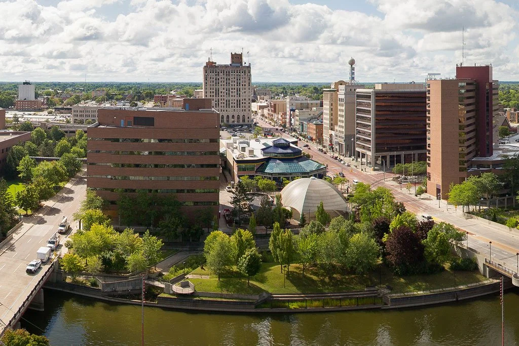 A view of a downtown cityscape with several tall buildings, green trees, a river in the foreground, and a partly cloudy sky.