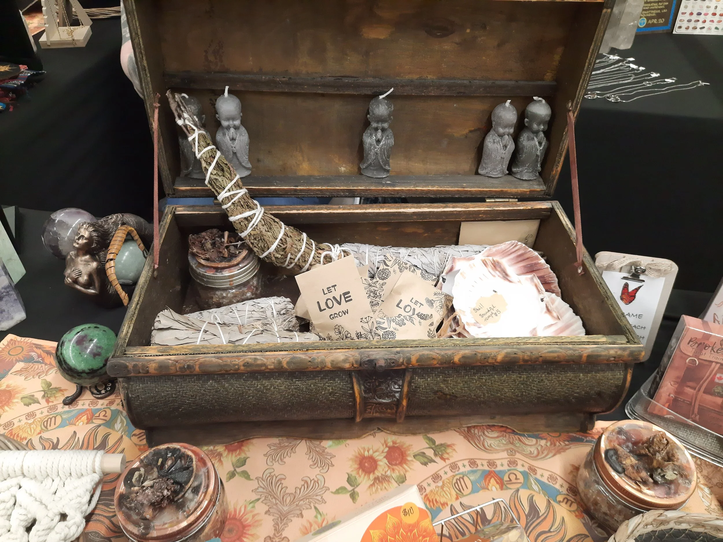 An open wooden box containing small statues, shells, cards with motivational phrases, and herbs or incense sticks. The box is on a table with various other items around it.
