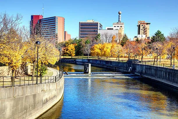 Cityscape with a river running through a park, surrounded by trees with yellow and green leaves, and tall buildings in the background under a clear blue sky.