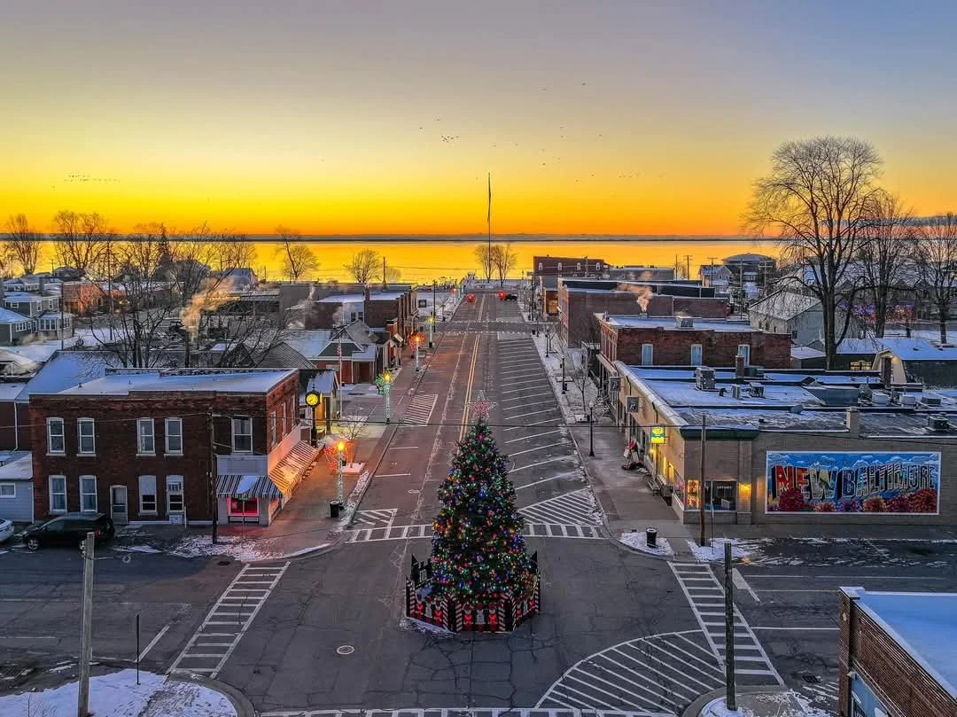 A small town square decorated for Christmas with a large decorated Christmas tree in the center, surrounded by snow, with brick buildings on either side, streetlights, and a sunset or sunrise over a body of water in the background.
