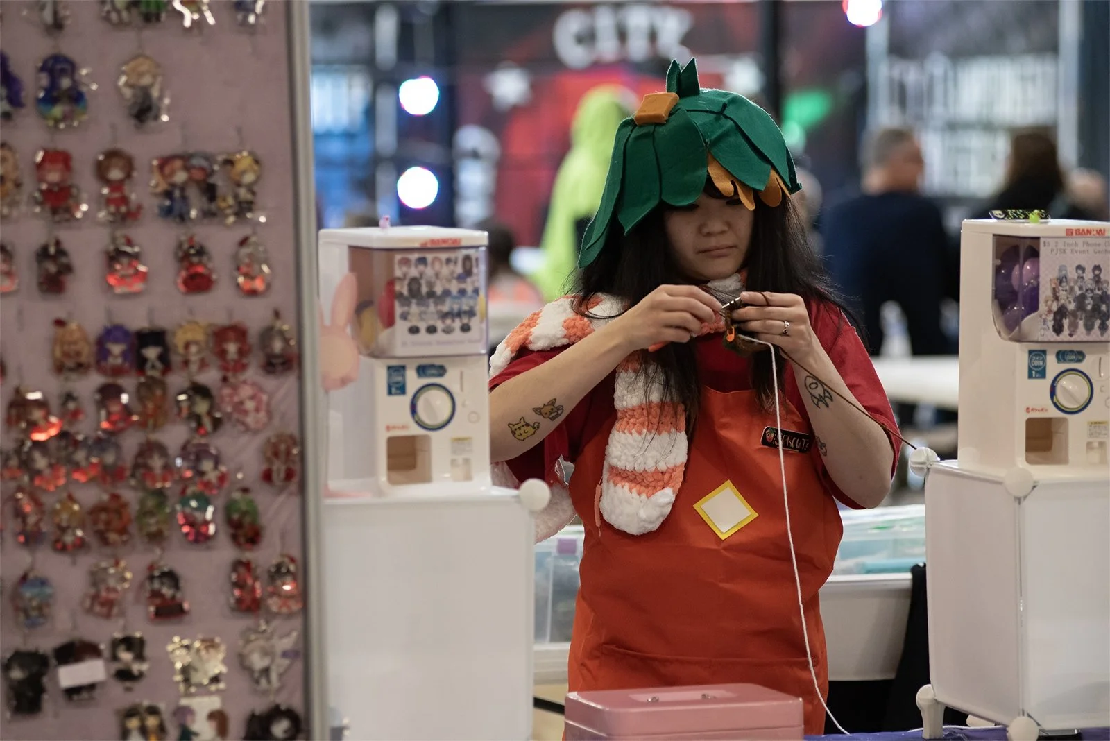 A woman wearing a green pumpkin hat and a red apron, working at a booth with small toys and souvenirs, surrounded by blurred shoppers and colorful lights.