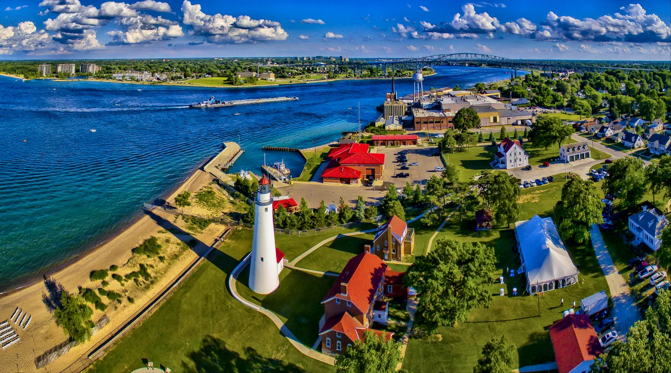 Aerial view of a riverfront town featuring a lighthouse, beach, residential houses, a parking lot, a church, and industrial buildings with a bridge in the background under a partly cloudy sky.