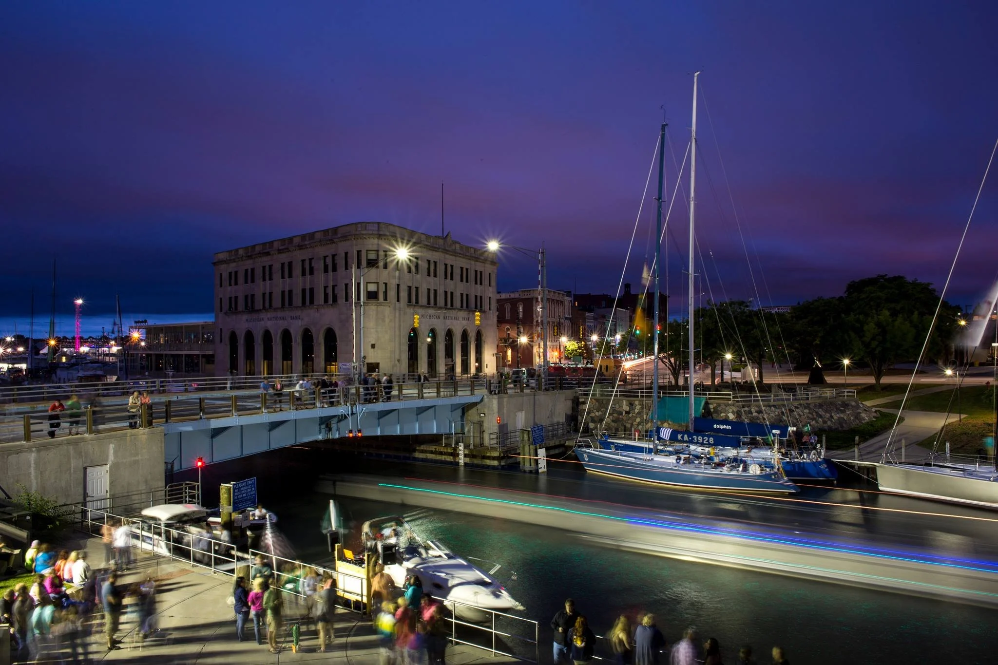 Night scene at a marina with boats, a historic building, and people walking along the waterfront