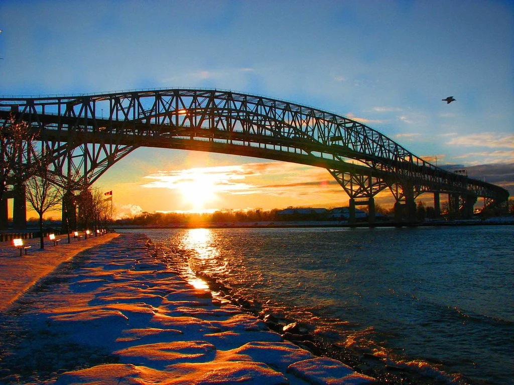 Bridge over a river at sunset with snow on the ground and a bird flying in the sky.