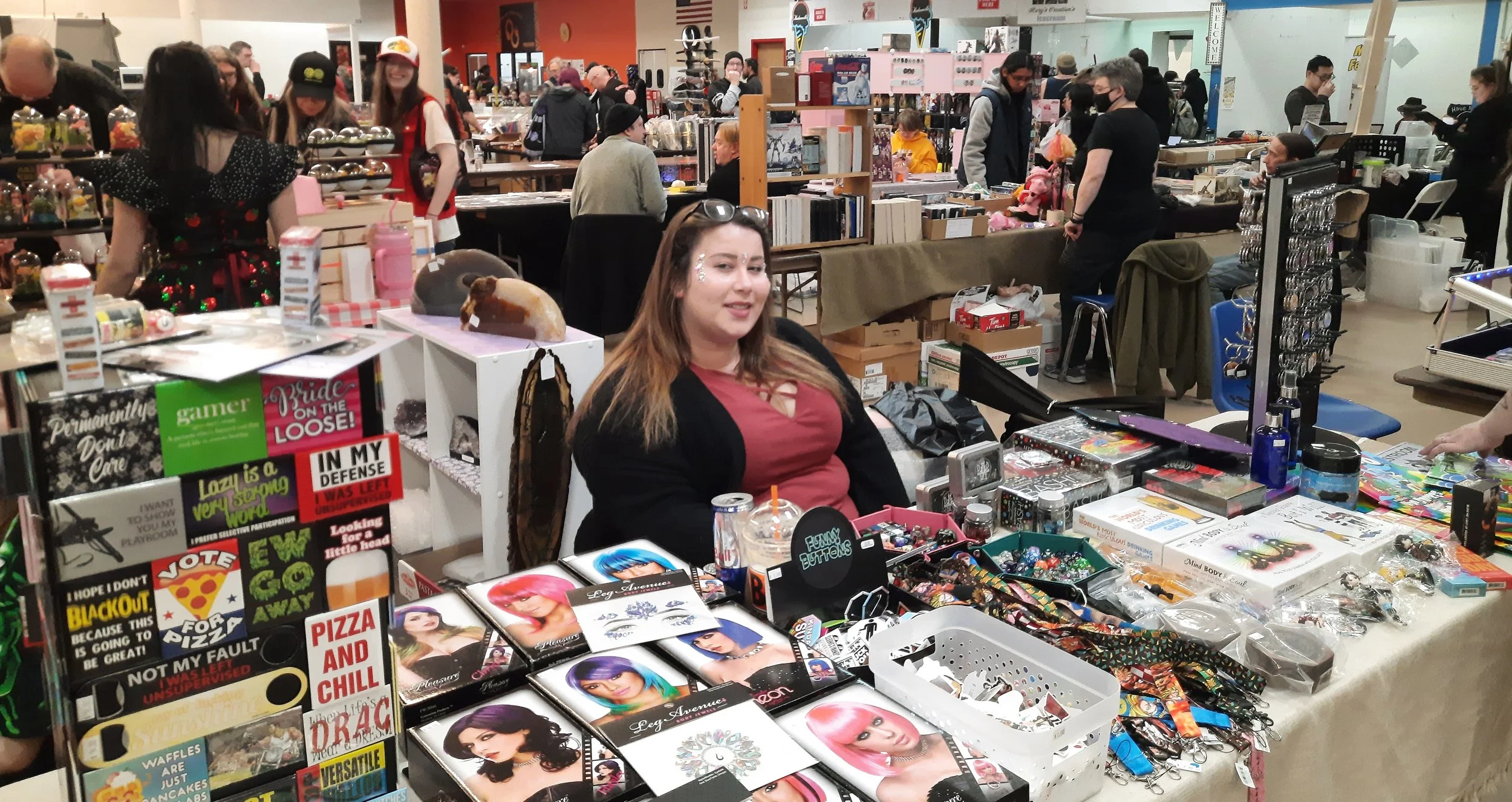 Female vendor sitting at a booth with jewelry, accessories, and decorative items during a market event, with shoppers browsing in the background.