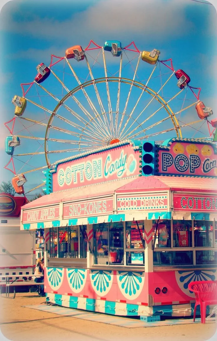 A colorful fairground stand with signs for cotton candy, snow cones, and cold drinks in front of a Ferris wheel at an amusement park.