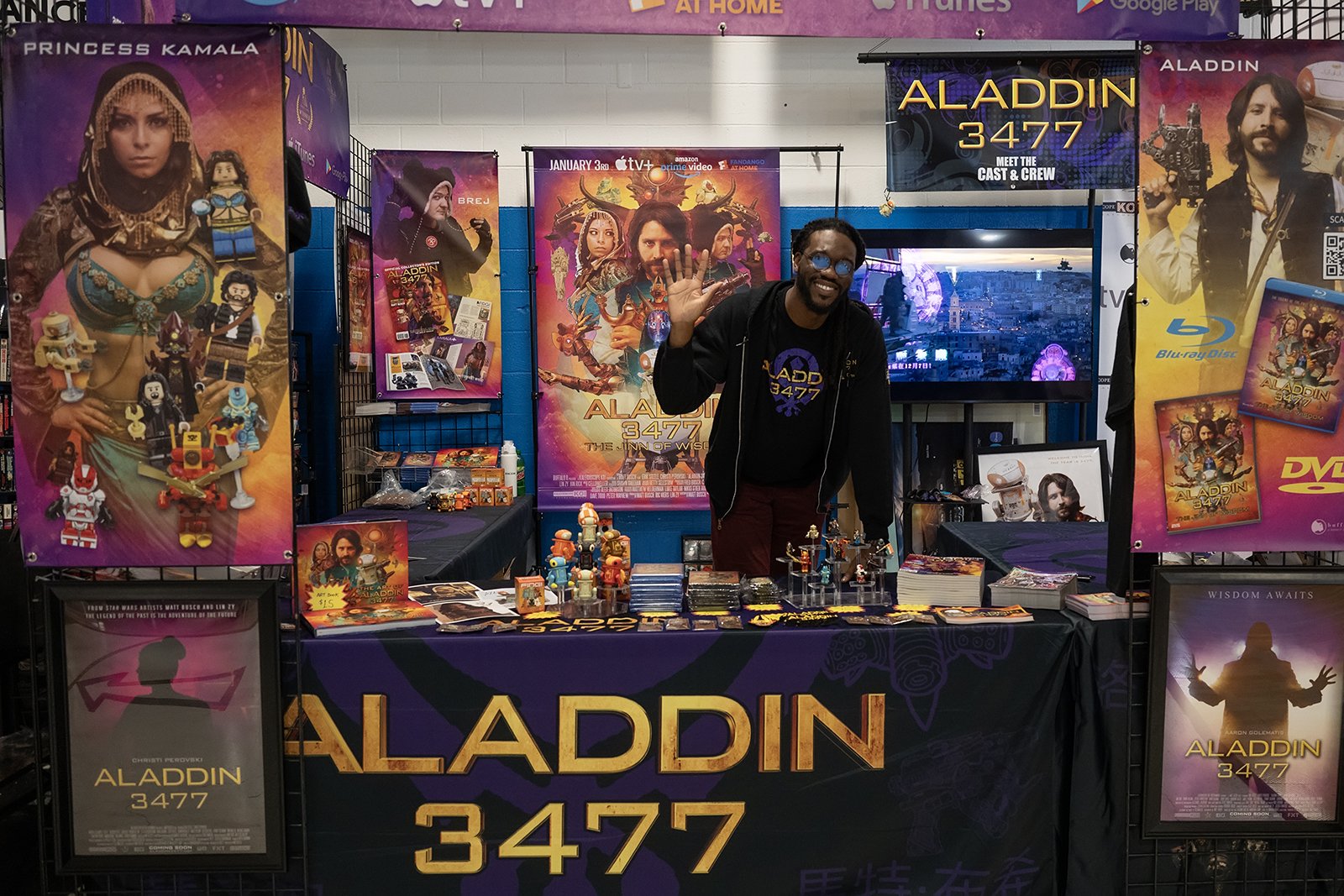 A man standing behind a table at a promotional booth for the movie 'Aladdin 3477.' The booth features colorful banners and posters, including one with a woman dressed as a princess and various LEGO figures, as well as TV screens displaying scenes from the film. The man is smiling and waving, wearing glasses and a black shirt with the 'Aladdin 3477' logo.
