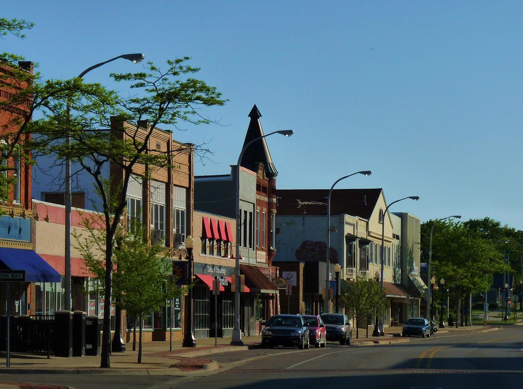 A small downtown street with shops, trees, parked cars, and streetlights, under a clear blue sky.
