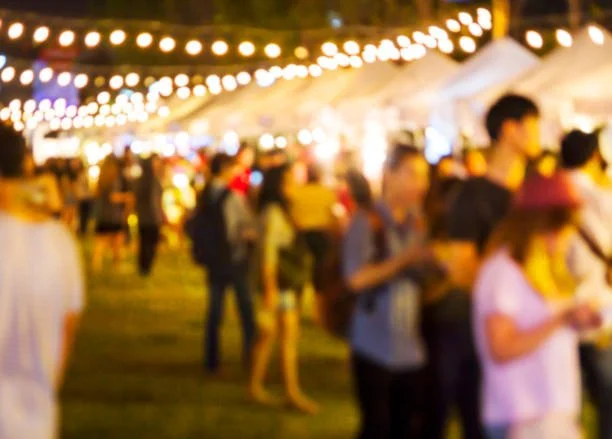 Blurred image of a nighttime outdoor event with string lights, tents, and people walking and socializing.