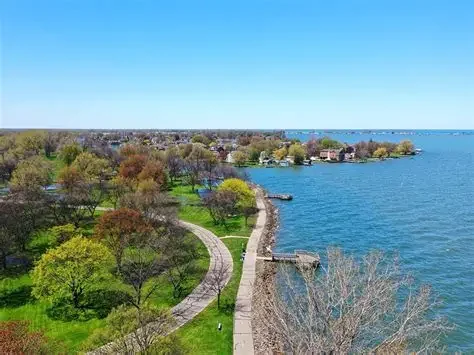 A scenic coastal park with a walking path, trees, and a view of the waterfront under a clear blue sky.