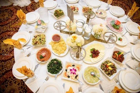 A round dining table set with various Middle Eastern dishes, including hummus, salad, and dips. White plates, utensils, and yellow napkins are arranged around the table. Decorative flowers and candles are in the center.
