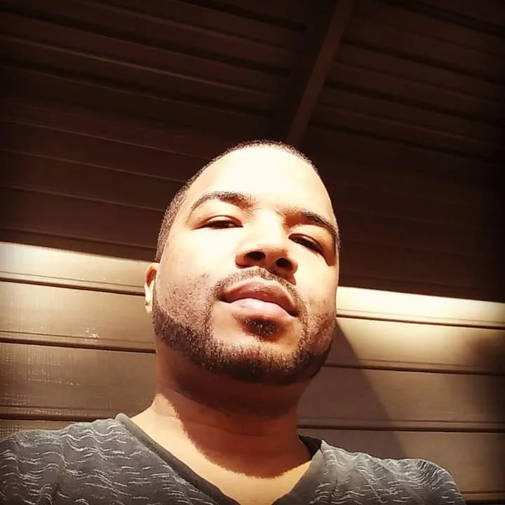 Man with a beard looking at the camera, wearing a dark shirt, against a wooden background.