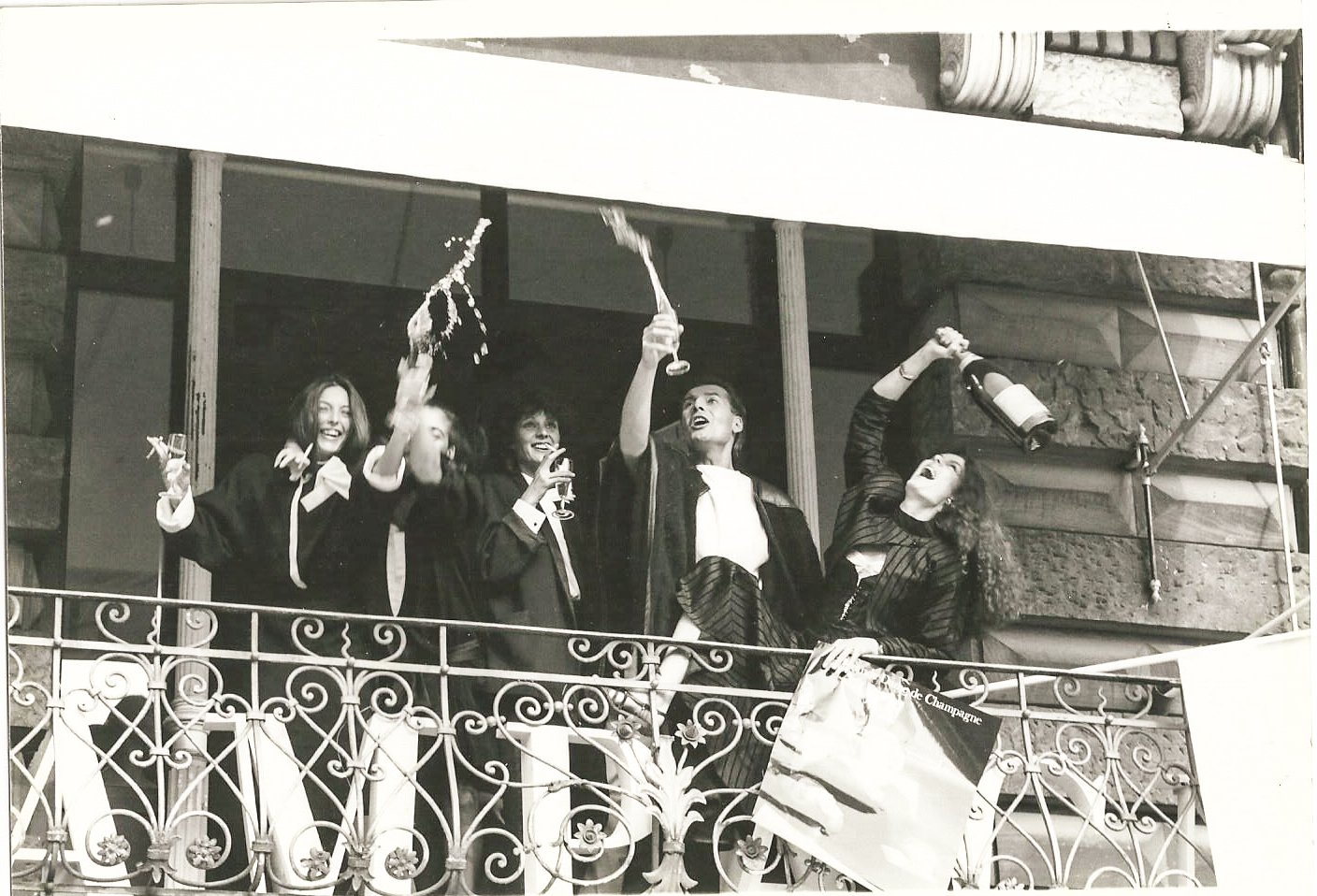 Fünf Menschen auf einem Balkon, die feiern und mit Sektgläsern und Flaschen anstoßen, mit Lachen und gehobenen Armen. Der Balkon hat ein schmiedeeisernes Geländer. Schwarz-weiß Foto.