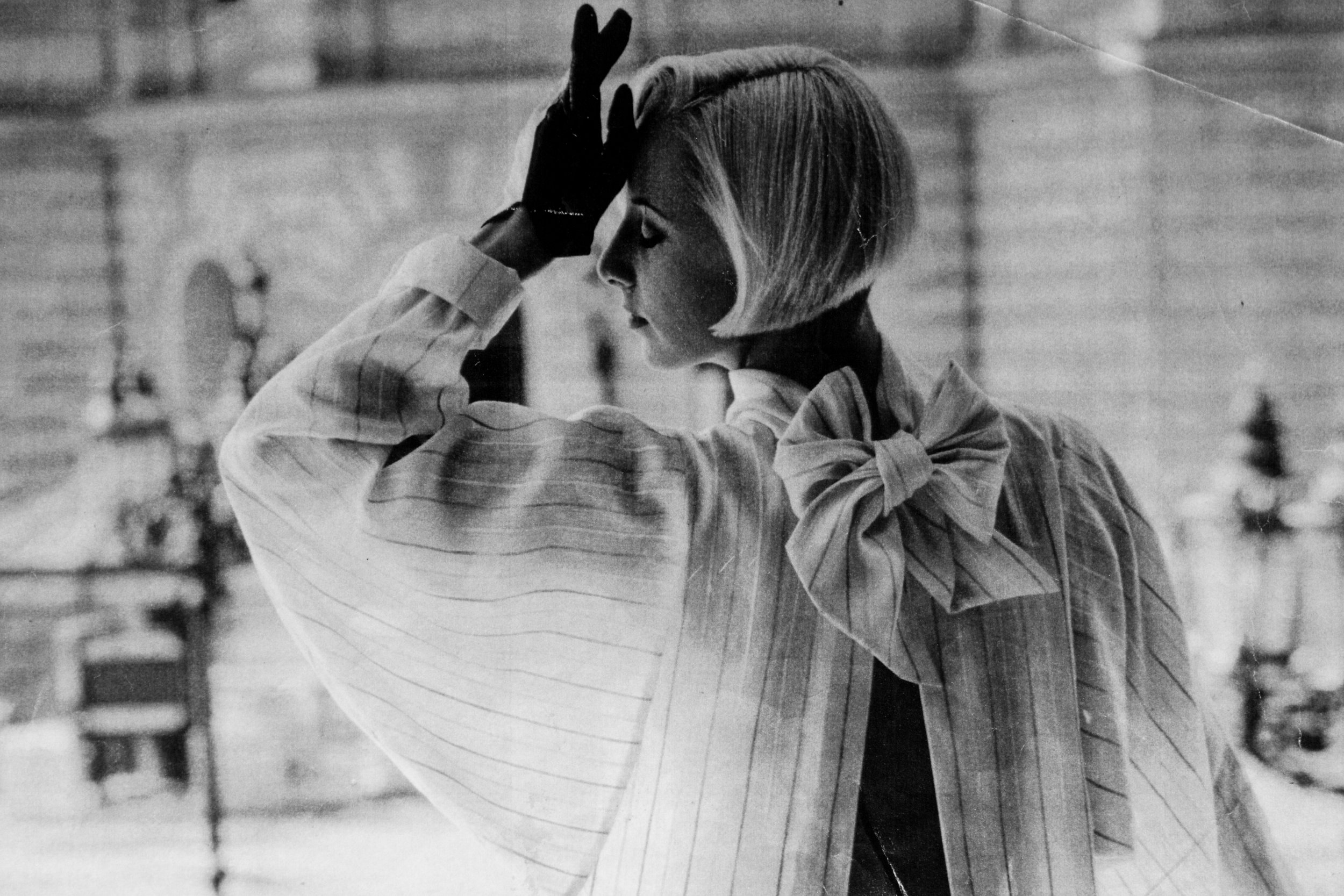Schwarz-weiß-Fotografie einer Frau von hinten mit kurzem Haar, eleganter gestreifter Bluse und großer Schleife am Hals. Sie hebt eine behandschuhte Hand an die Stirn. Im Hintergrund ist unscharfe Architektur zu sehen.