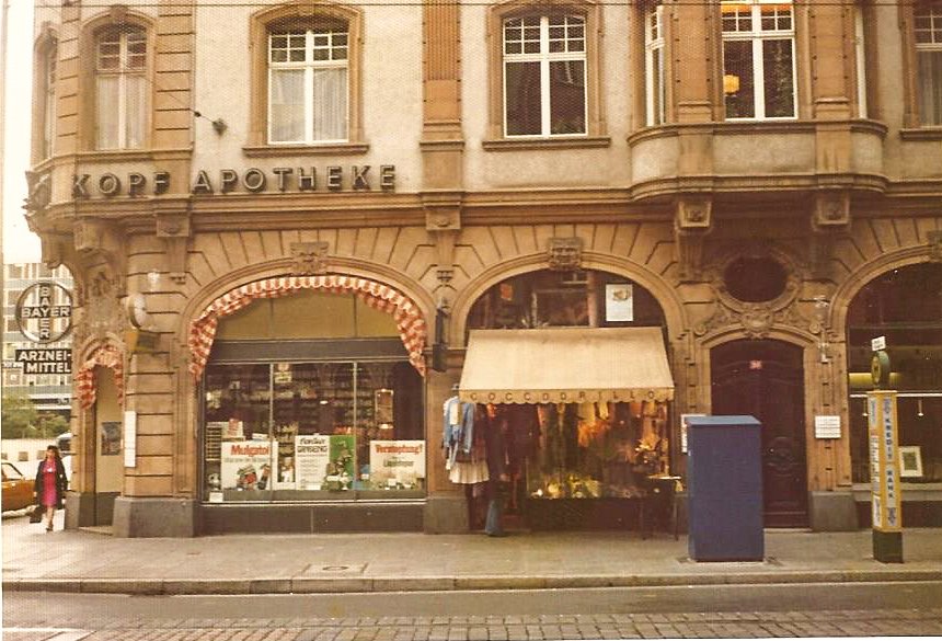 Historisches Gebäude mit einer Apotheke im Erdgeschoss, 'Kopf Apotheke' genannt. Die Fassade ist mit Schaufenstern dekoriert. Ein Laden mit Markise und Kleidungsstücken ist zu sehen. Eine Person geht auf dem Bürgersteig vorbei und ein blaues Objekt s