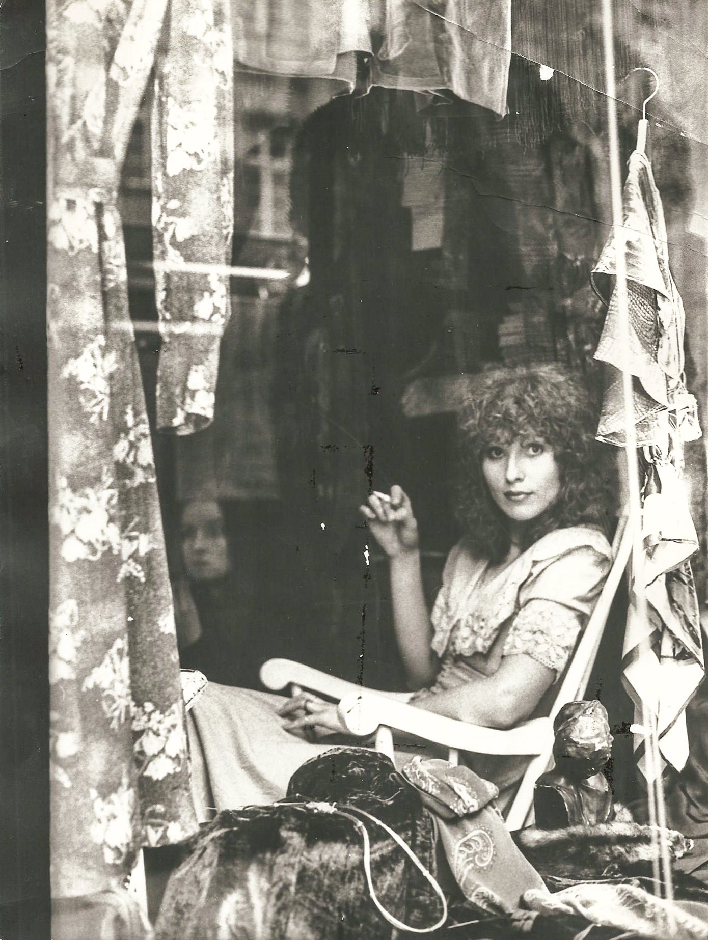 Schwarzweißfoto einer Frau mit lockigem Haar, die in einem Schaukelstuhl sitzt. Um sie herum sind Stoffe und Kleidung zu sehen. Die Szene wirkt vintage und intim.