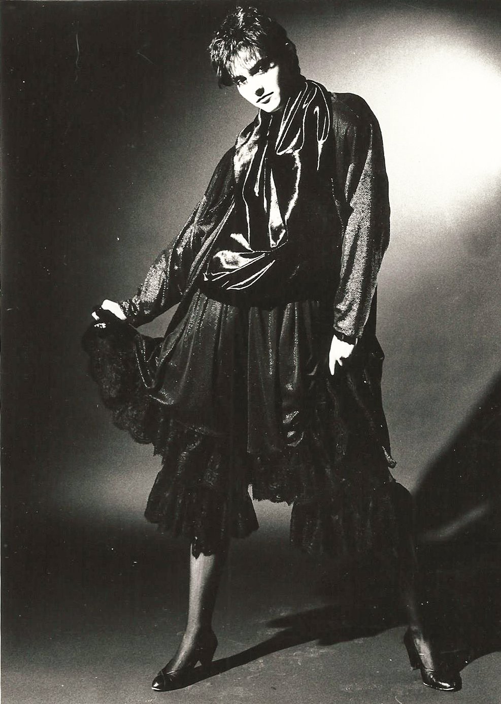 Schwarz-Weiß-Fotografie einer Person in eleganter schwarzer Mode, mit einer glänzenden Bluse und einem voluminösen Rock, in einer Studioszene. Die Pose ist expressiv und künstlerisch.
