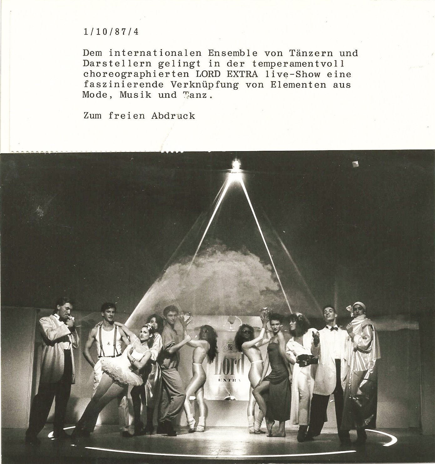 Schwarz-Weiß-Foto einer Bühnenshow mit Tänzern und Darstellern in verschiedenen Posen. Im Hintergrund ist ein Banner mit der Aufschrift "Lord Extra" sichtbar. Oben befindet sich ein Text über die Show.
