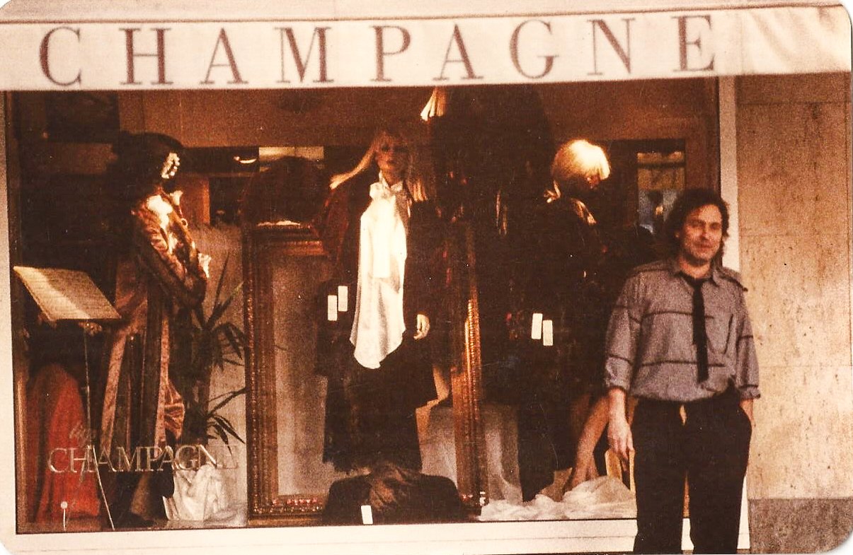 Schaufenster eines Geschäfts mit dem Schild 'Champagne'. In der Auslage stehen Schaufensterpuppen in historischer Kleidung. Rechts steht eine Person in einem grauen Hemd mit einer schwarzen Krawatte und schwarzen Hosen.