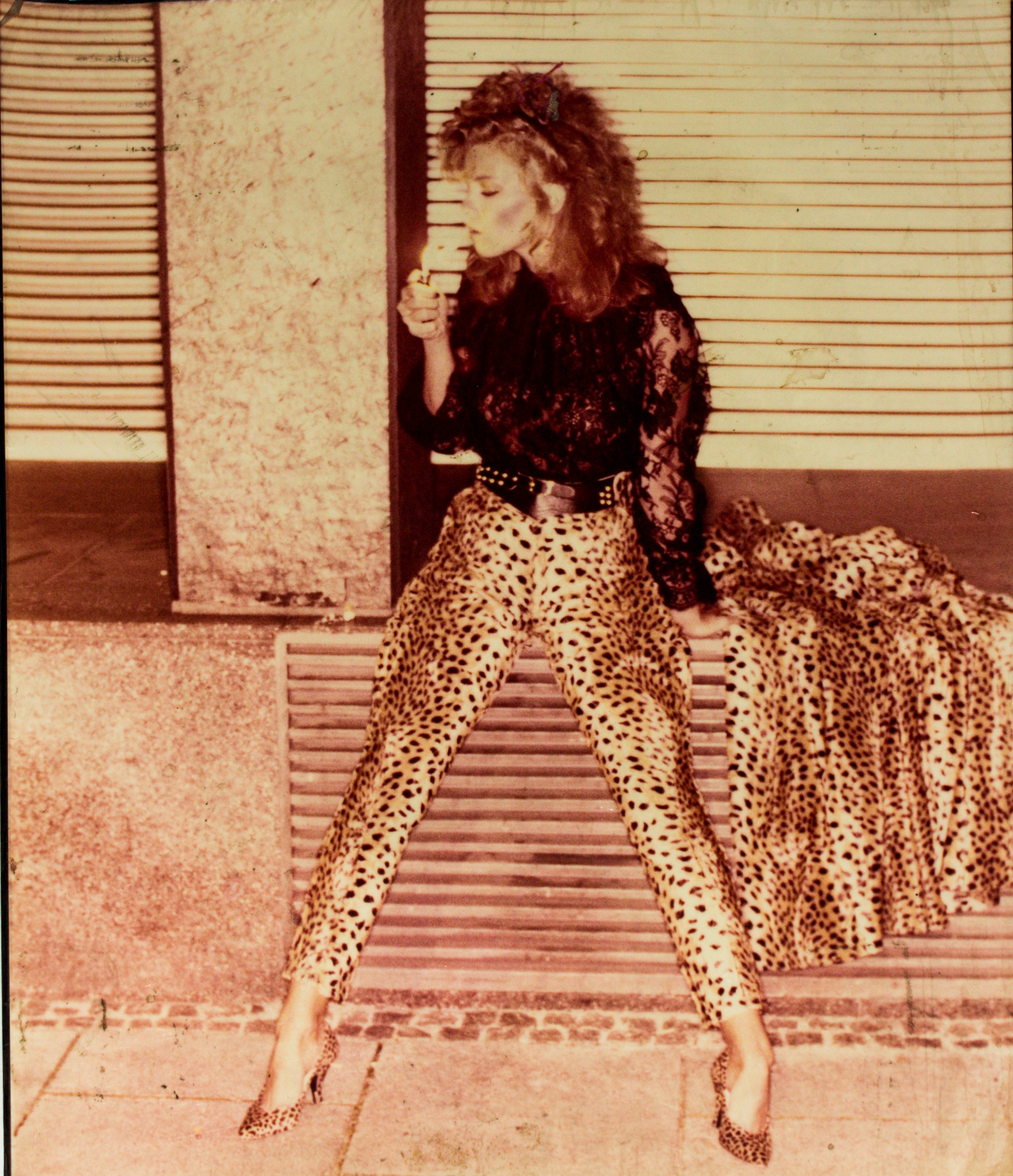 Frau in Leopardenmusterhose und Spitzenbluse sitzt auf einer Bank und raucht. Sie trägt High Heels und hat lockiges Haar.