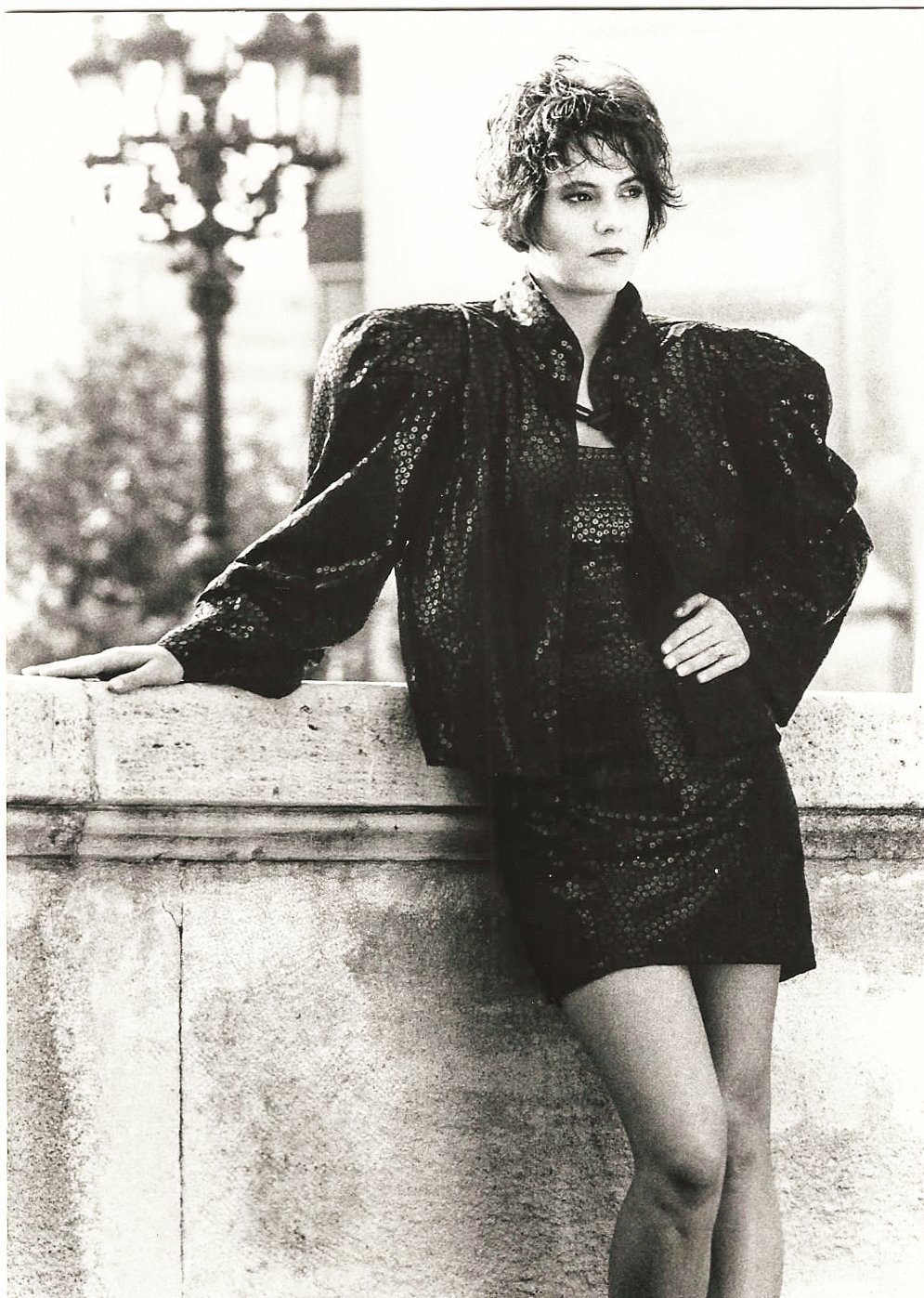 Schwarz-weiß-Foto einer Person in einem eleganten Outfit mit Puffärmeln, stehend an einer Steinmauer, im Hintergrund verschwommene Stadtansicht.