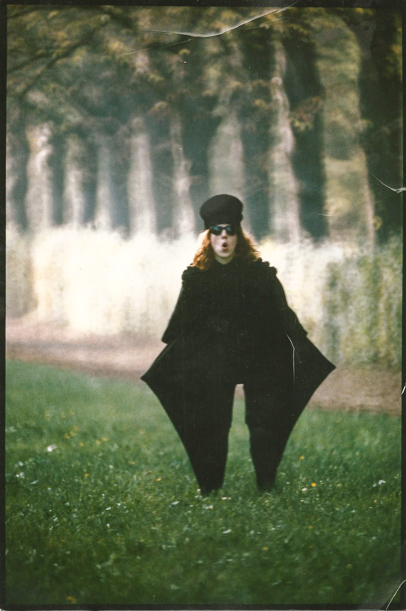 Person in schwarzer Kleidung im Freien, trägt Sonnenbrille und Hut, umgeben von Bäumen und Wiese.