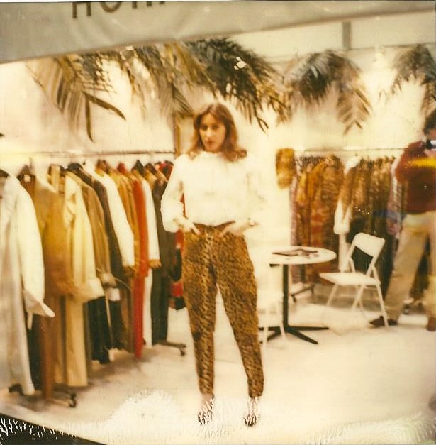 Eine Person steht in einem Bekleidungsgeschäft mit Leopardenhose vor einem Ständer voller Kleidung. Im Hintergrund sind Palmenblätter und ein Tisch mit einem Stuhl zu sehen. Die Atmosphäre wirkt retro.