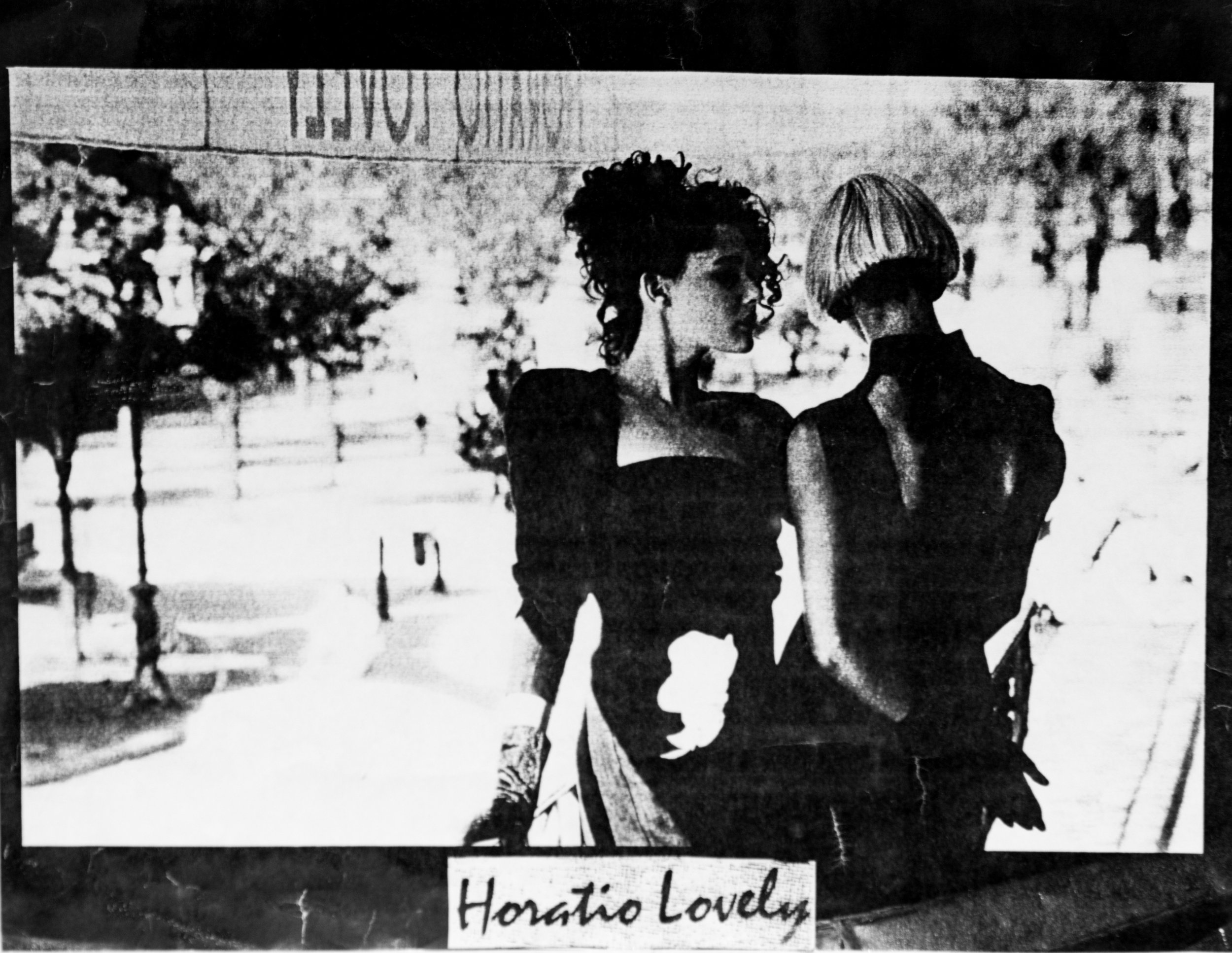 Zwei Frauen in eleganter Kleidung stehen auf einem Balkon. Der Hintergrund zeigt einen unscharfen Park mit Bäumen. Unten im Bild steht "Horatio Lovely" in eleganter Schrift auf einem Schild.