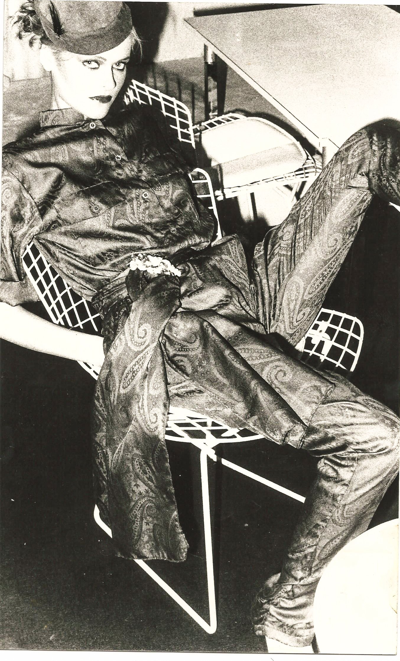 Person in einem stilvollen, gemusterten Anzug sitzt lässig auf einem Drahtstuhl, mit intensivem Blick und einem Hut, in einem monochromen Foto.