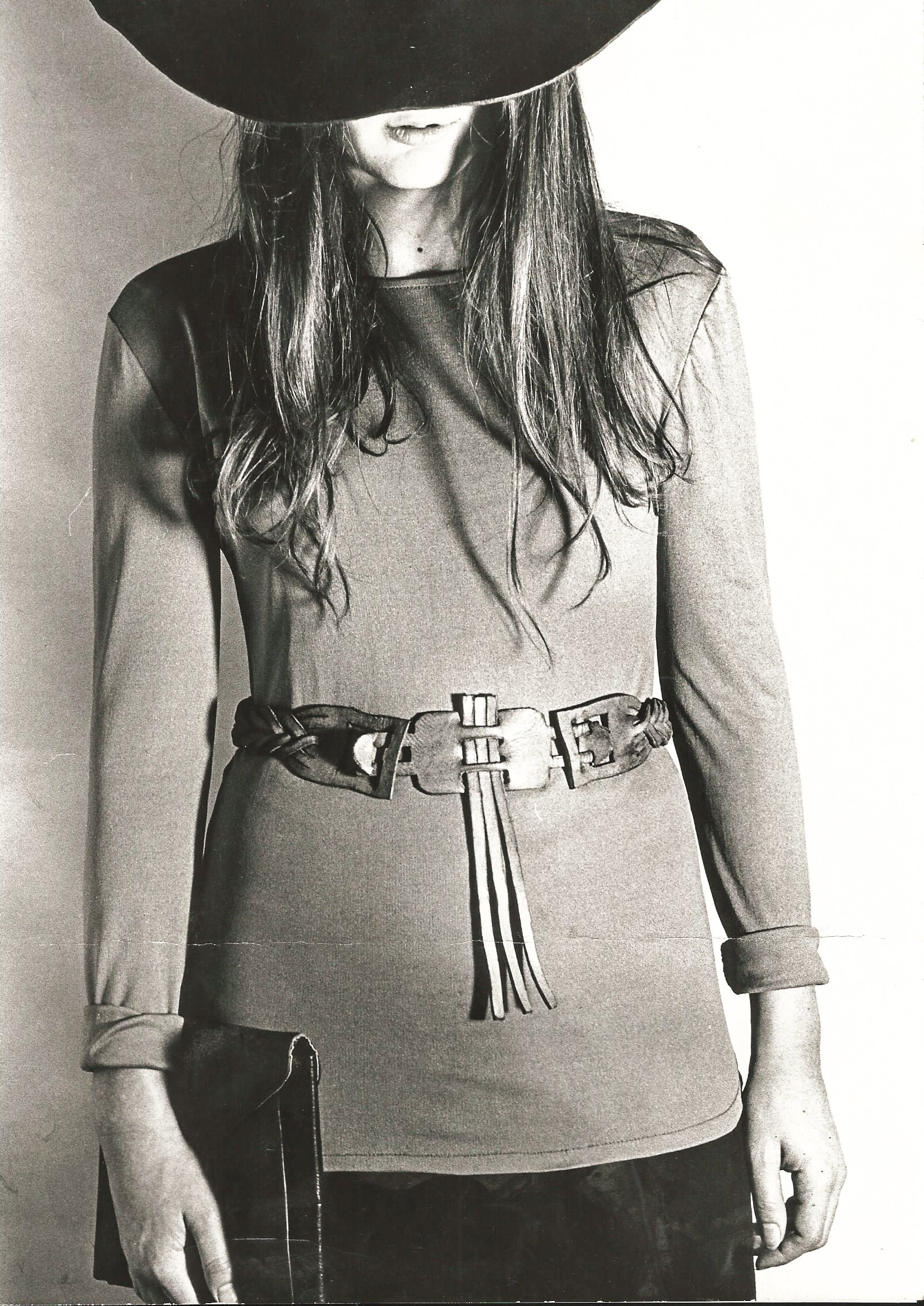Schwarz-Weiß-Foto einer Frau, deren Gesicht von einem großen Hut verdeckt wird, trägt ein langärmliges Oberteil mit Gürtel und hält eine Clutch.