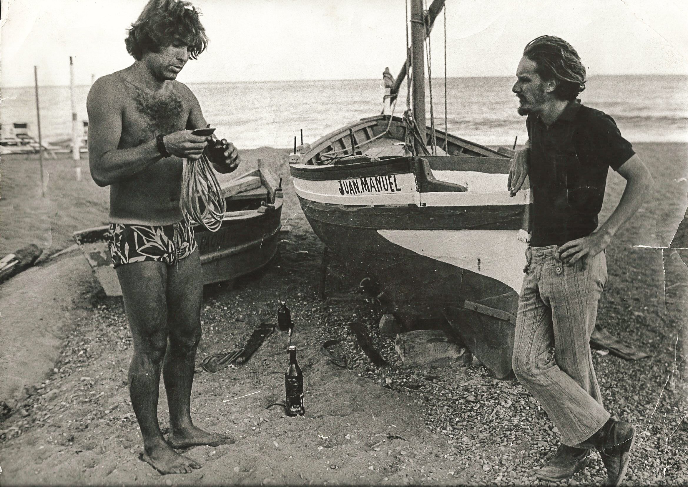 Zwei Männer stehen am Strand neben Fischerbooten. Einer der Männer trägt eine Badehose und hält Seil, der andere steht lässig daneben. Im Hintergrund ist das Meer zu sehen und eine der Boote trägt den Namen "JUAN MANUEL". Es gibt auch ein paar Flasch