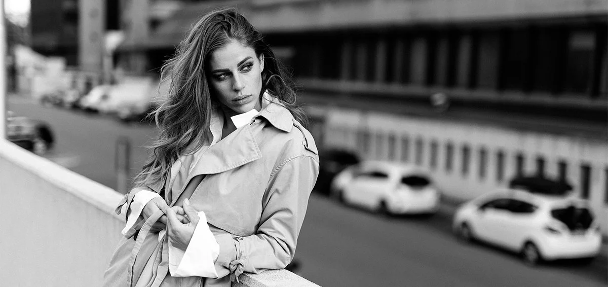 Donna con cappotto su un ponte, sfondo di città, immagine in bianco e nero.