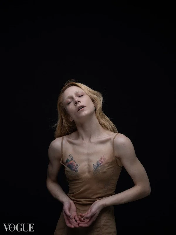 Persona con capelli biondi che indossa un abito beige, con tatuaggi visibili sulle spalle, su sfondo nero.