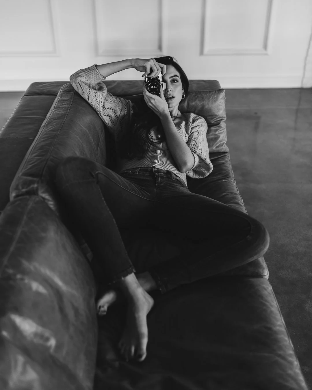 Donna sdraiata su un divano, tiene una macchina fotografica e guarda l'obiettivo, foto in bianco e nero.