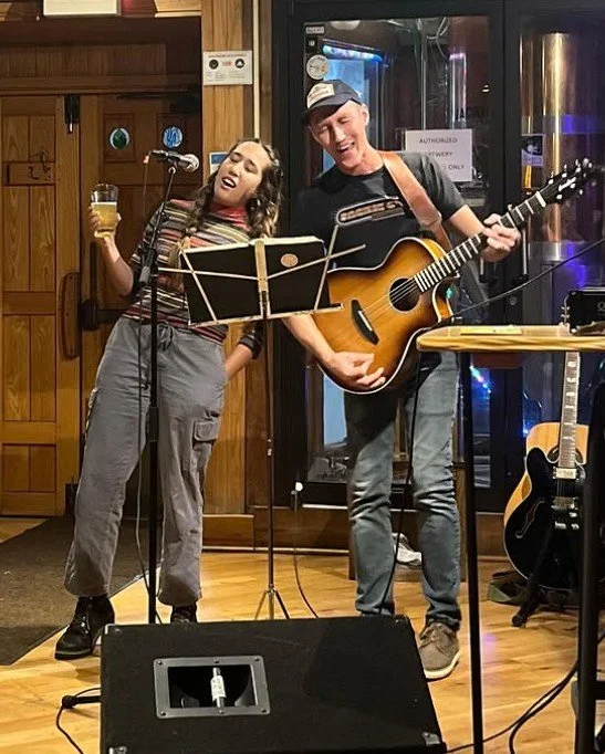 Two people performing on stage, one singing into a microphone and the other playing a guitar. The woman holds a drink, and both appear to be enjoying the performance. Musical equipment and seating can be seen in the background.