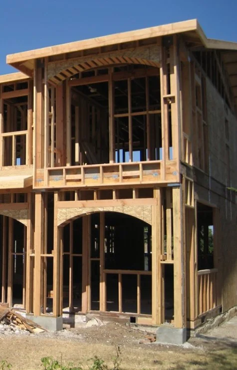 Wood framing of a two-story house under construction with visible open windows and doorways, set against a clear blue sky.