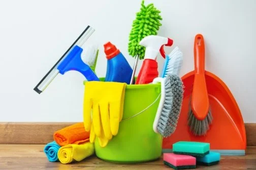Cleaning supplies including spray bottles, scrub brushes, sponges, rubber gloves, a dustpan, and cloths in front of a plain background.