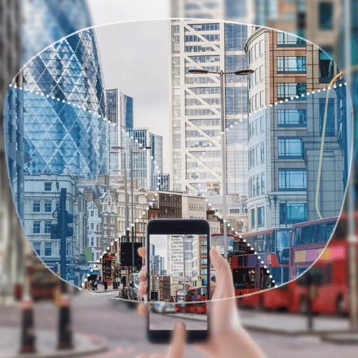 A person taking a photo of a city street scene with a smartphone, showing tall modern buildings and red double-decker buses.