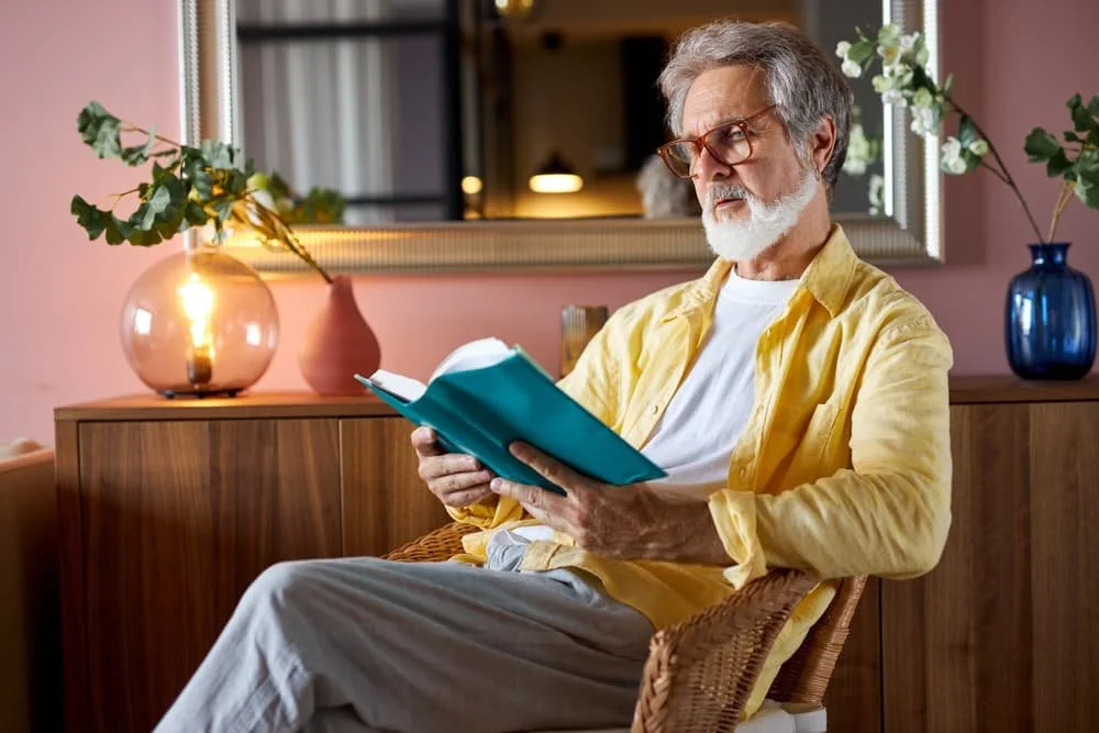 An older man with glasses and a white beard sitting in a wicker chair, reading a teal book indoors, with a wooden sideboard, decorative lamps, and vases with flowers behind him.