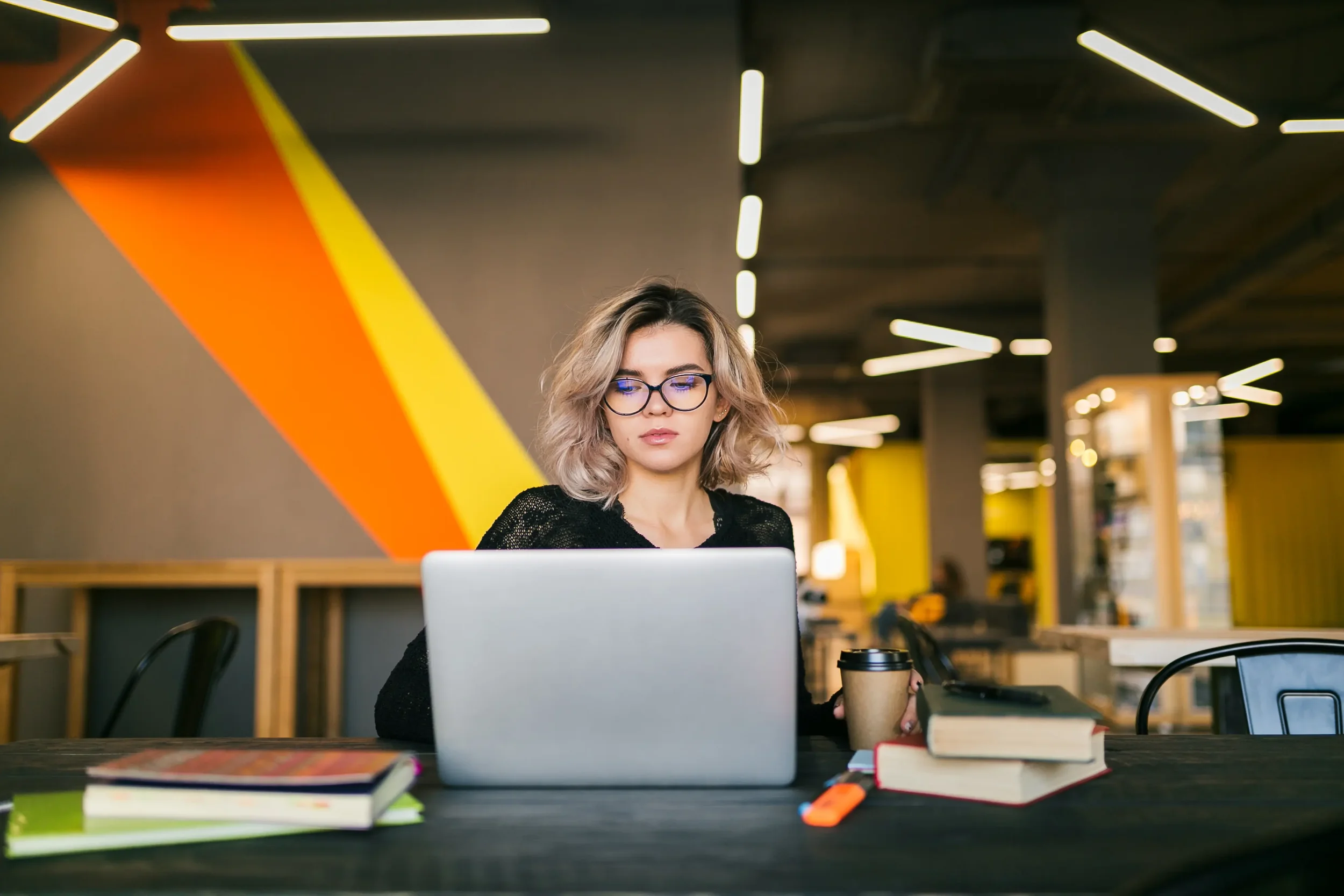 A young woman with curly blonde hair and glasses working on a laptop at a desk in a modern, brightly lit cafe or co-working space, with books, a coffee cup, and a highlighter on the table.