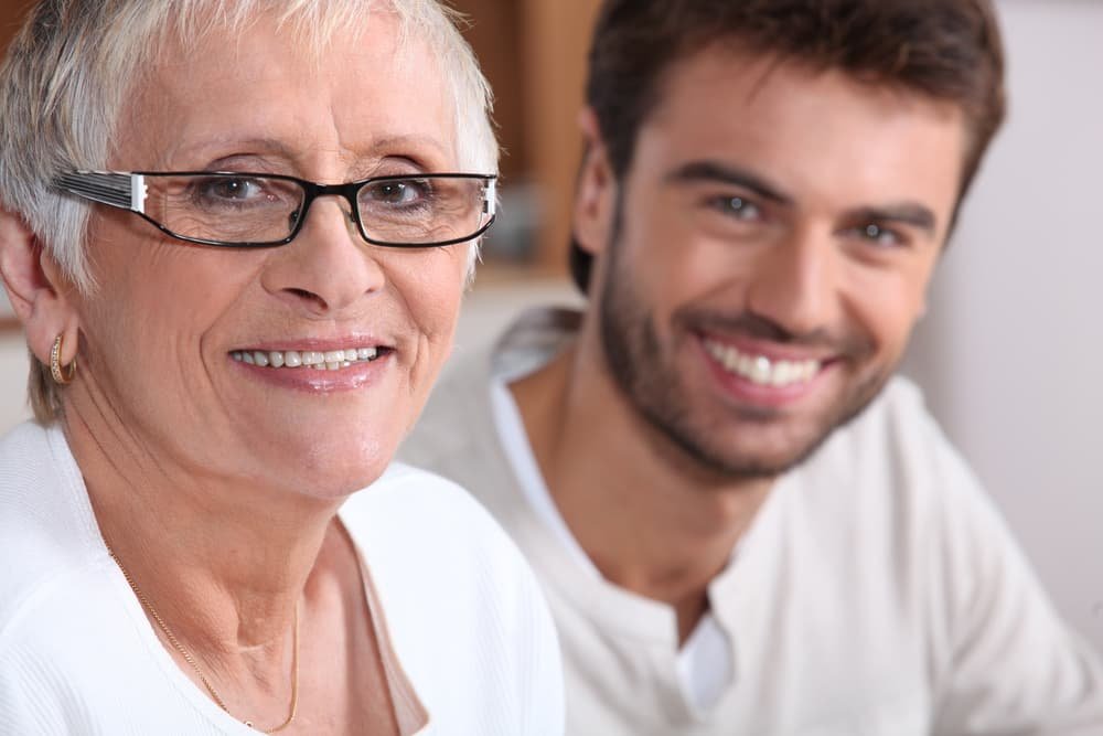 An elderly woman with glasses and a young man, both smiling, in a close-up portrait.