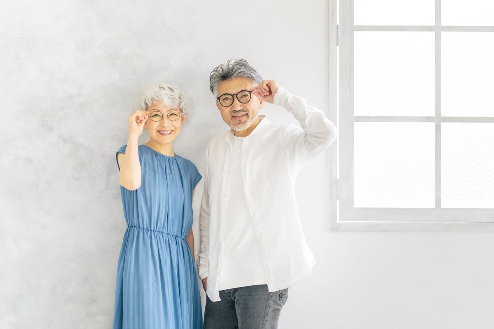 A mature Asian couple wearing glasses, smiling, and adjusting their glasses while standing in a bright room with a window.