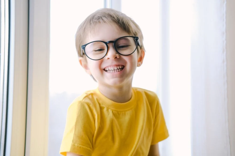 A smiling young boy with light brown hair wearing glasses and a yellow t-shirt, standing near a window with sunlight behind him.