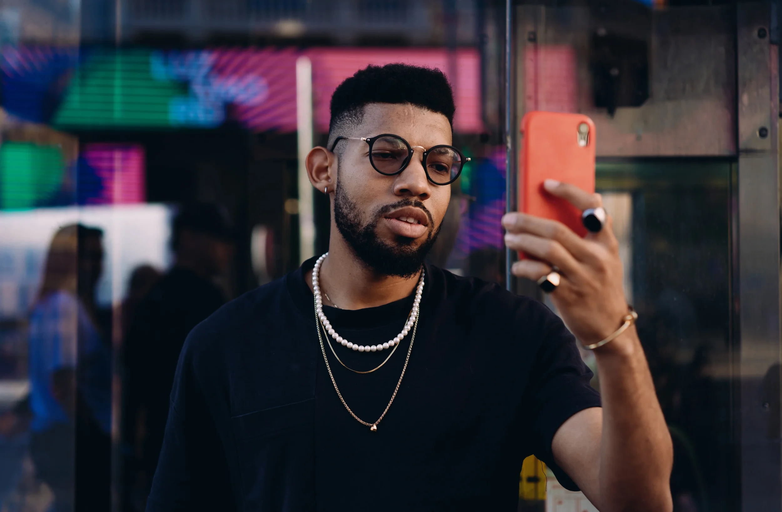 Man with short hair, sunglasses, and jewelry taking a selfie with a pink phone in an urban setting with neon lights.