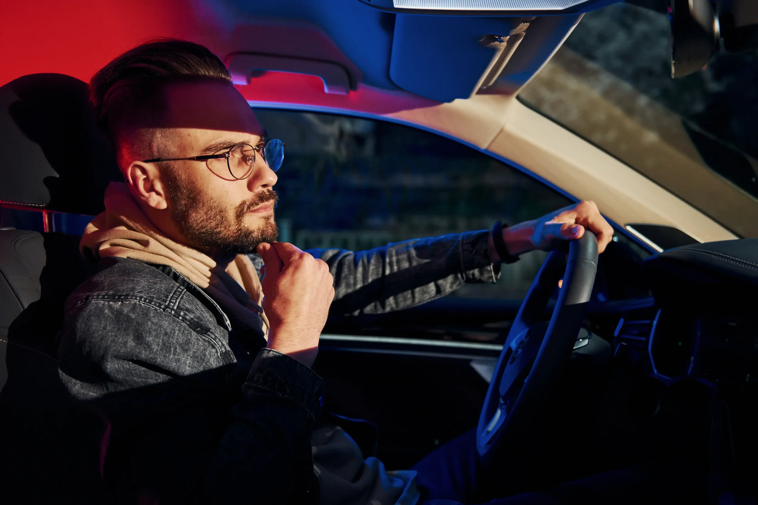 A man with glasses and a beard sitting in the driver's seat of a car at night, looking thoughtful with one hand on his chin and the other on the steering wheel.