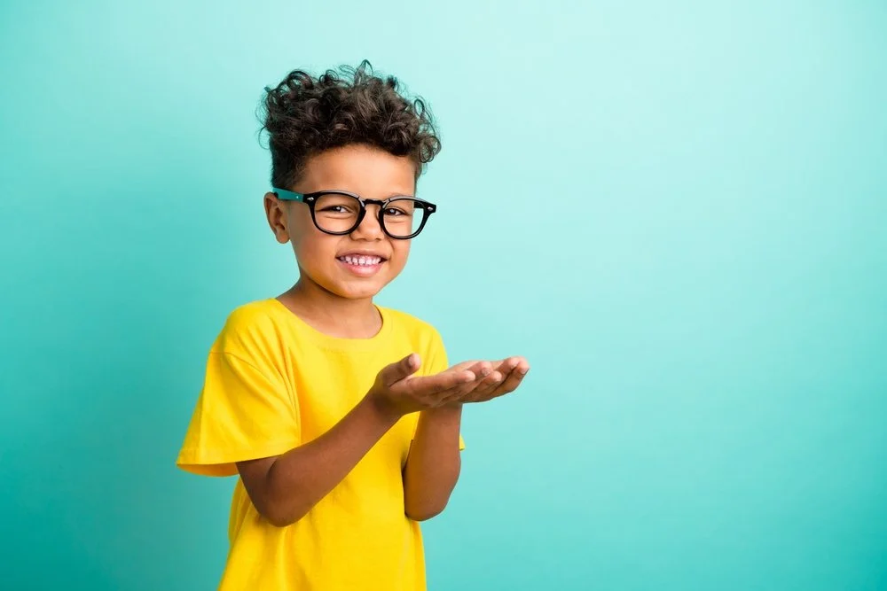 A young boy with curly hair, glasses, and a yellow t-shirt smiling and holding his hands together against a light blue background.