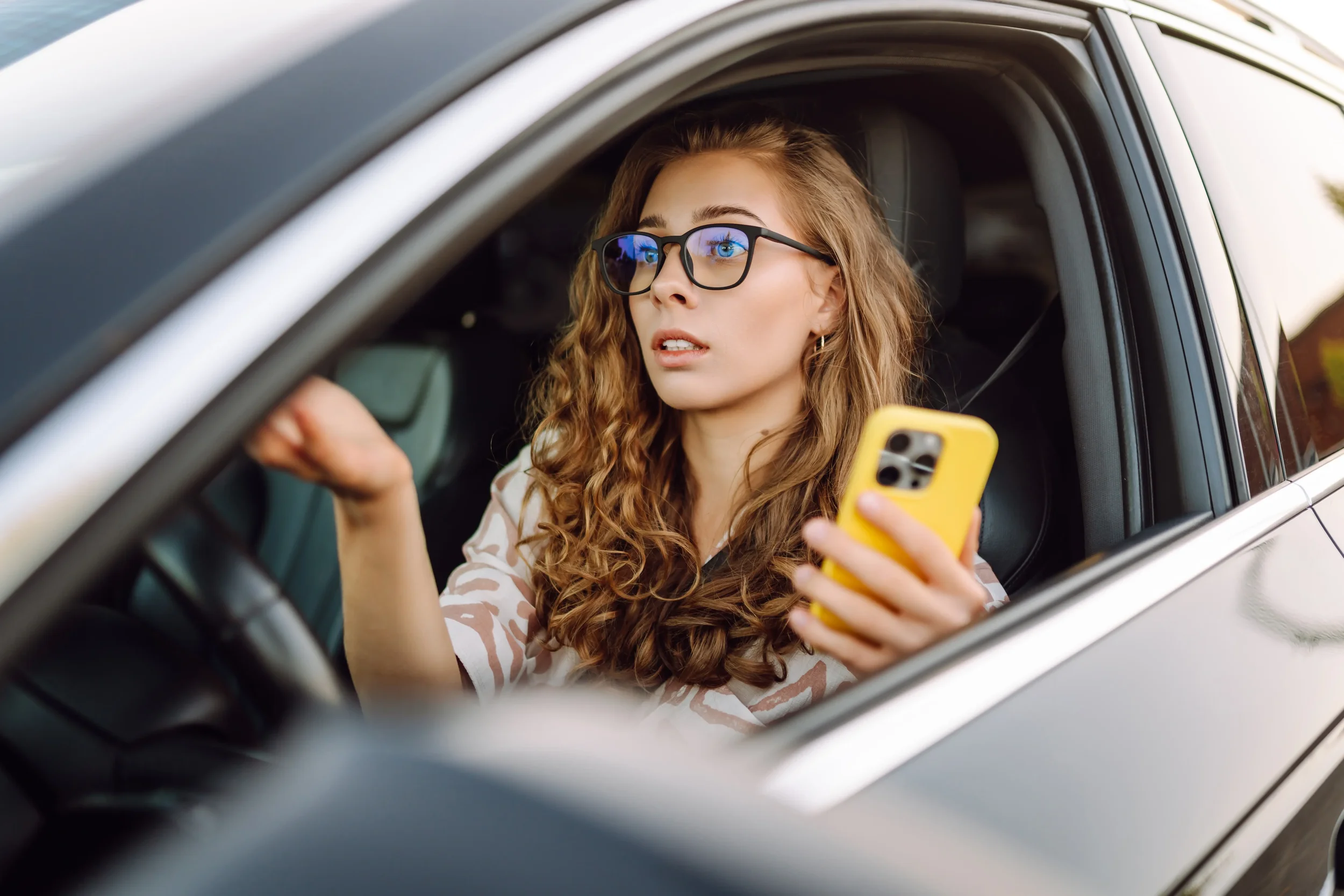 A woman with long curly hair, wearing glasses, sitting in the driver's seat of a car, holding a yellow phone, looking out the window with a focused expression.