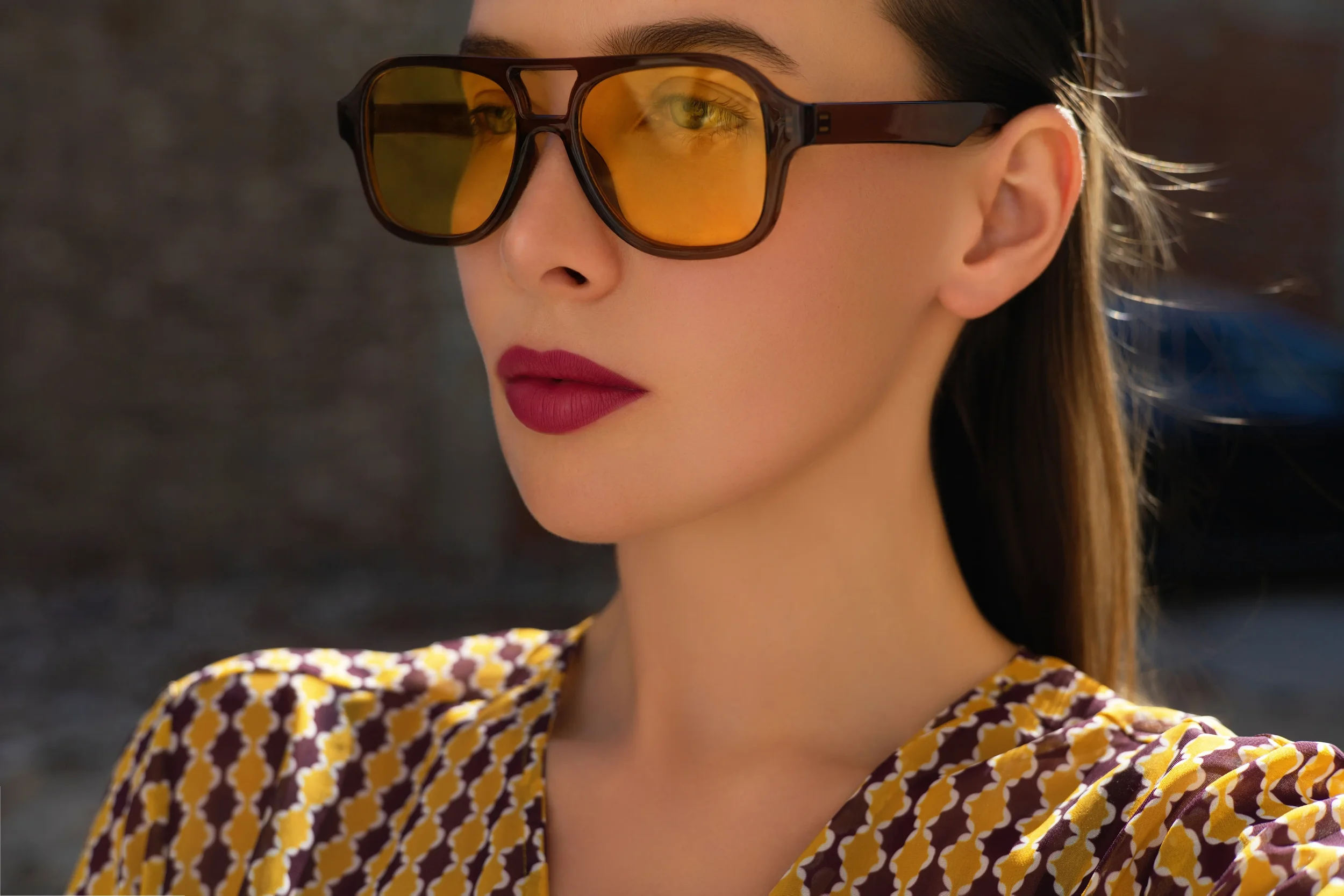 A woman with brown hair, wearing large sunglasses with yellow-tinted lenses, red lipstick, and a patterned yellow and brown dress.